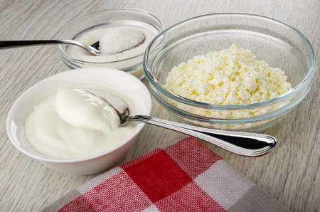 Teaspoon in bowl with sugar, spoon in white bowl with sour cream, transparent glass bowl with grainy cottage cheese, checkered napkin on wooden tableの写真素材