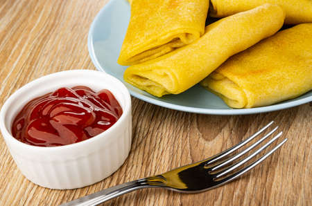 White bowl with ketchup, pancake rolls with filling in blue plate, fork on brown wooden tableの写真素材
