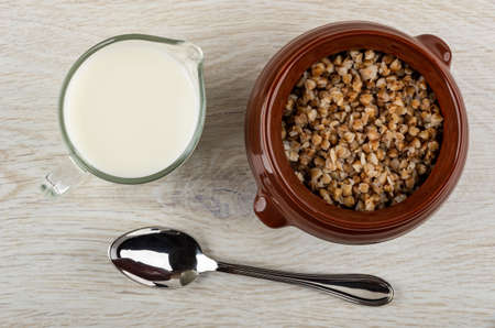 Glass jug with milk, ceramic brown pot with cooked buckwheat porridge, spoon on wooden table. Top viewの写真素材