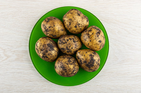 Few raw dirty unpeeled potatoes in green plate on wooden table. Top viewの写真素材