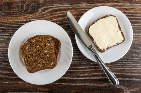 Few slices of rye bread with sunflower seeds in glass plate, table knife and sandwich with butter in white saucer on dark wooden table. Top viewの写真素材
