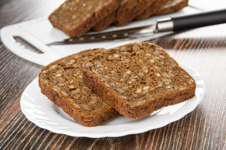 Slices of rye bread with sunflower seeds, kitchen knife on plastic cutting board, white plate with...