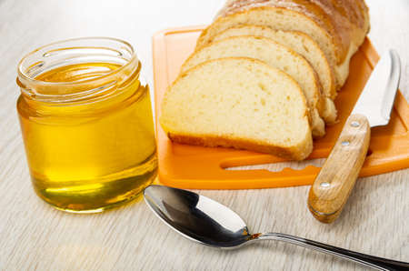 Opened transparent glass jar with liquid honey, slices of sweet bun, kitchen knife on cutting board, spoon on wooden tableの写真素材