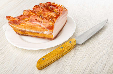 Piece of smoked brisket in vacuum packaging on white plate, kitchen knife on wooden table