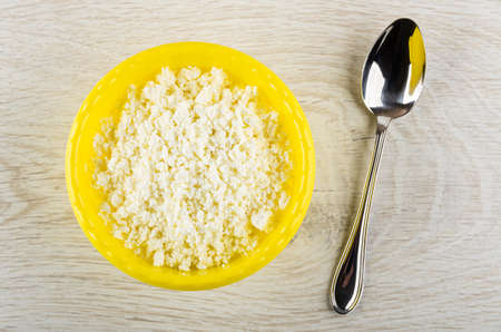 Grainy cottage cheese in yellow plastic bowl, metallic spoon on wooden table. Top view