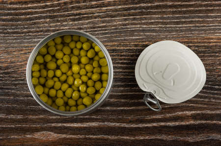 Opened metallic jar with canned green peas, lid on dark wooden table. Top view