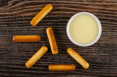 Scattered small bread sticks, bowl with condensed milk on dark wooden table. Top view