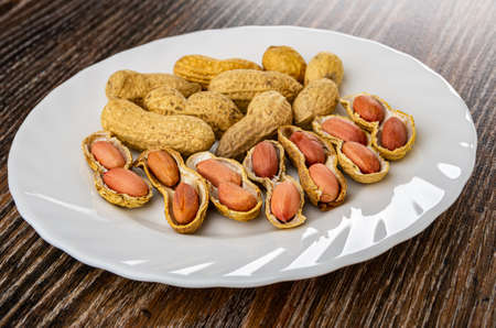 Fried peanuts in peel, peeled groundnuts in white glass plate on dark wooden table