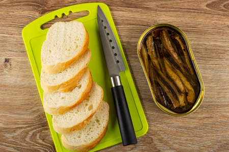 Slice of wheat bread, kitchen knife on green cutting board, opened metallic jar with canned sprats on wooden table. Top viewの写真素材