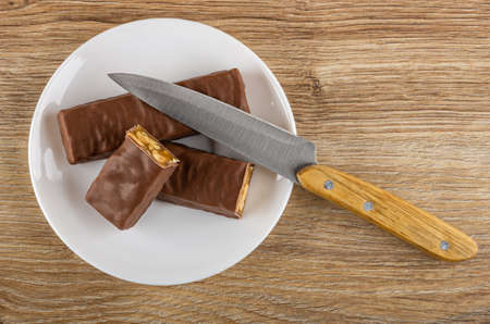 Chocolate bar with peanut and caramel, halves of candy, kitchen knife in white glass plate on wooden table. Top viewの写真素材