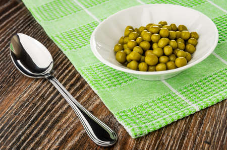 Metallic spoon, white bowl with canned green peas on green napkin on dark wooden tableの写真素材