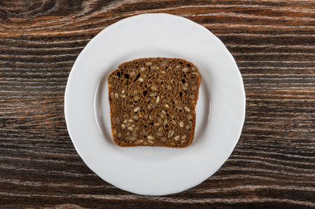 Slice of rye bread with sunflower seeds in white plate on dark wooden table. Top view