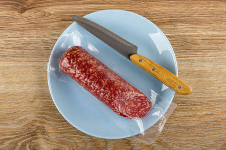 Smoked sausage in vacuum packaging, kitchen knife in blue glass plate on wooden table. Top view