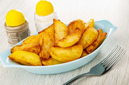 Pepper shaker and salt shaker, blue oval plate with slices of fried potato, fork on wooden table