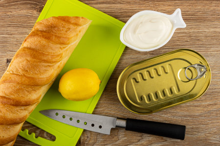 Fresh yellow lemon and wheat baguette on green cutting board, bowl with mayonnaise, closed jar with canned fish, kitchen knife on wooden table. Top viewの写真素材