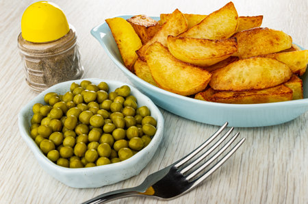 Pepper shaker, bowl with canned green peas, blue oval plate with slices of fried potato, fork on wooden tableの写真素材
