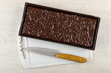 Chocolate wafer cake in plastic box, kitchen knife on cutting board on wooden table. Top view