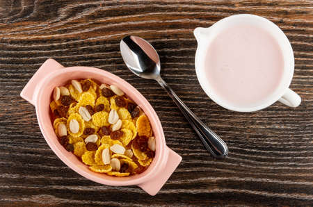 Pink oval bowl with corn flakes, raisin, peanut, teaspoon, pitcher with fruit yogurt on dark wooden...