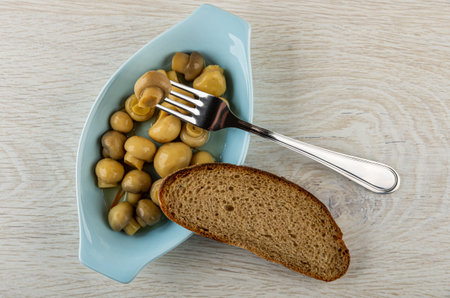 Mushroom strung on fork, slice of bread on blue oval plate with marinated champignons on wooden table. Top viewの写真素材
