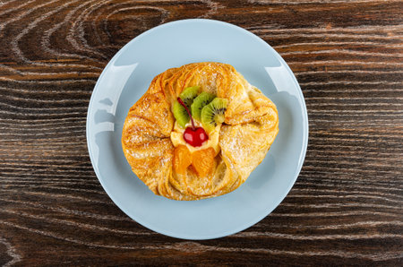 Danish bun with kiwi, tangerine and cherry in blue plate on dark wooden table. Top view