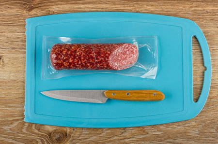 Smoked sausage in vacuum packaging, kitchen knife on blue plastic cutting board on wooden table. Top viewの写真素材
