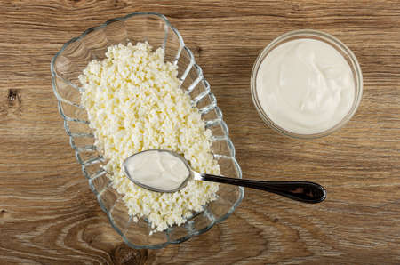 Spoon with sour cream in transparent glass bowl with grainy cottage cheese, bowl with sour cream on...