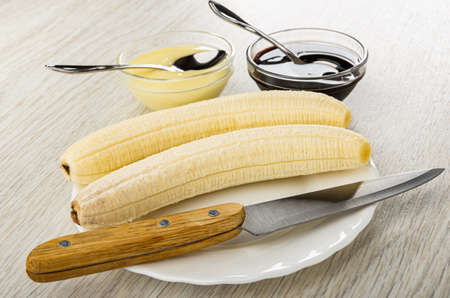 Transparent bowls with condensed milk and chocolate sauce two peeled bananas, kitchen knife in white plate on wooden tableの写真素材