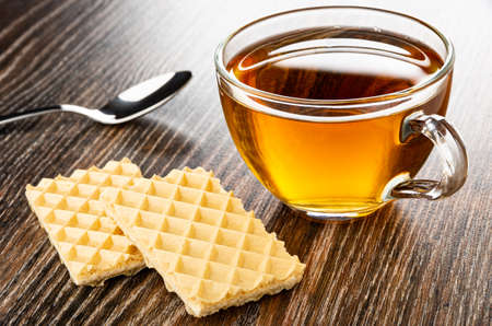 Teaspoon, transparent glass cup with hot tea, wafers on dark wooden table