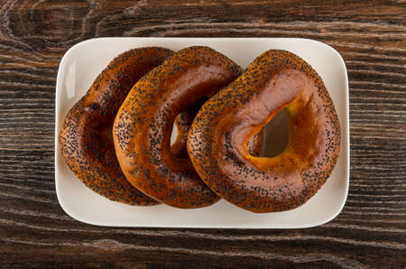 Baked bread rings with poppy seeds in white glass plate on dark wooden table. Top viewの写真素材