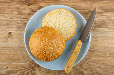 Halves of burger bun with sesame seeds and kitchen knife in blue glass plate on wooden table. Top...