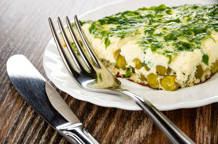 Table knife, fork on white plate with slice of omelet with chopped parsley and green peas on dark wooden tableの写真素材