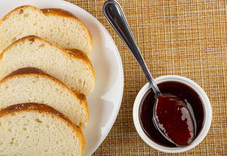 Slices of sweet bun in white glass plate teaspoon in white glass bowl with strawberry jam on brown...