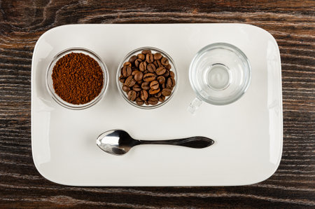 Transparent bowls with ground coffee and roasted coffee beans, empty coffee cup, spoon in white plate on dark wooden table. Top viewの写真素材