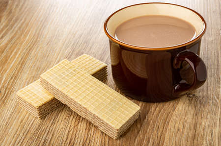 Two wafers, brown glass cup of cocoa with milk on wooden table