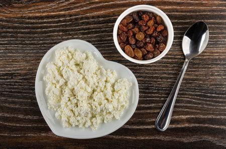 White saucer in heart shape with cottage cheese, glass bowl with raisin, teaspoon on dark wooden table. Top viewの写真素材