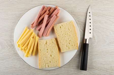 Piece of cheese, sausages, slices of wheat bread in white glass plate, kitchen knife on wooden...