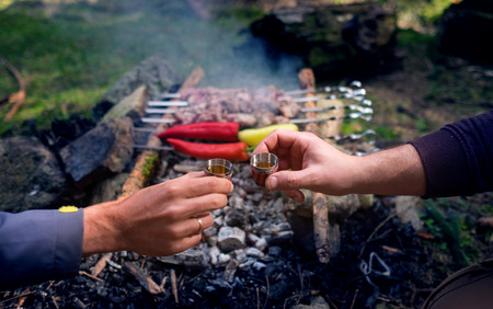 Two men hands clinking with bitter shots with grilled Shashlik background - the popular roasted on the Fire Meat meal.の写真素材