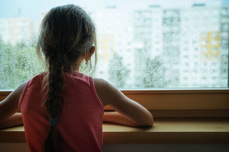 Little sad girl pensive looking through the window glass with a lot of raindrops. Sadness  and...