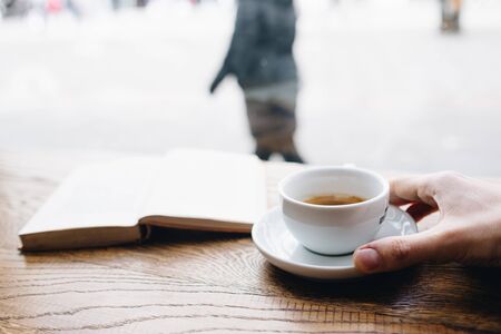 Human hand holding black coffee on saucer with vintage book on background on the wooden table next to big coffee shop window and looking to passerby street walkersの写真素材