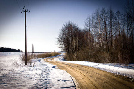 Landscape with the blue sky, road from gravel, snow, wood and bushes and a columnの写真素材