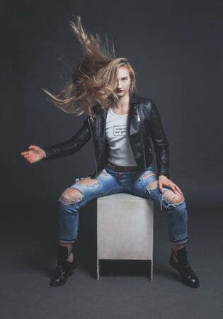 a young girl with long, flowing hair sits on a white cube on a gray background and makes a wave of her handの写真素材