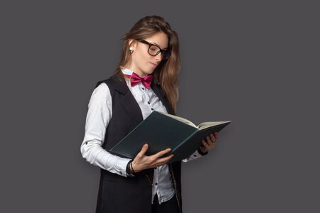 Young girl in white shirt and black vest with Burgundy bow tie and classic black pants office style and glasses looking into an open bookの写真素材
