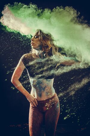 Beautiful young woman celebrating Holi paint fest with colorful powder covering her face and a cloud of dust. On black with rainbow bursts of colorの写真素材