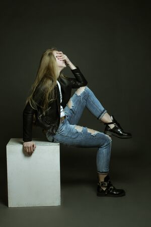 Studio portrait dark background laughing girl, covering her face with her hand. The model sits sideways on a white cube. She was wearing a white t-shirt, ripped jeans, and an even-cut leather jacket.の写真素材