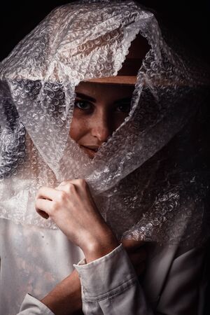 Studio portrait of a young girl in light brown, on a dark background, buttoning, using  wrap as a handkerchief over a hat. The concept of unnatural modern fabricsの写真素材
