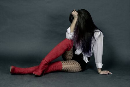 Brunette with dined hair in a white shirt and black shorts and red boots, sits on a grey background and covers her face the concept of depression,の写真素材
