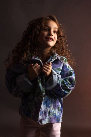 Studio portrait of a little girl with long curly hair, in a colorful jacket and light shorts. Smile...