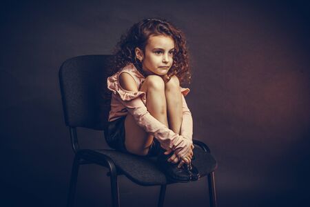 a little girl, with curls in a pink blouse and black shorts, sits on an office chair with her feet on her face, hurt, unhappy. Studio portraitの写真素材