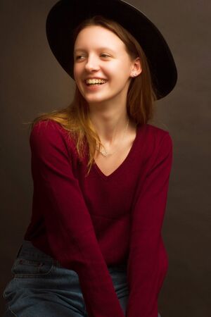 Studio portrait of a young blonde girl in a black hat, red blouse and blue jeans on a brown background.の写真素材
