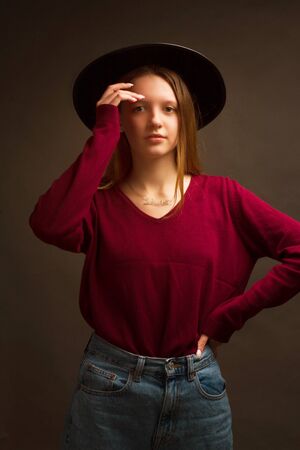 Studio portrait of a young blonde girl in a black hat, red blouse and blue jeans on a brown background.の写真素材